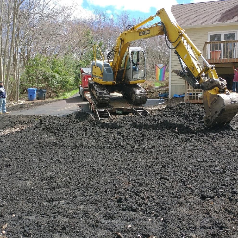 A yellow excavator from M Lato Excavating & Tree Care Service diligently moves soil in a residential yard, with a dump truck nearby and a house in the background.