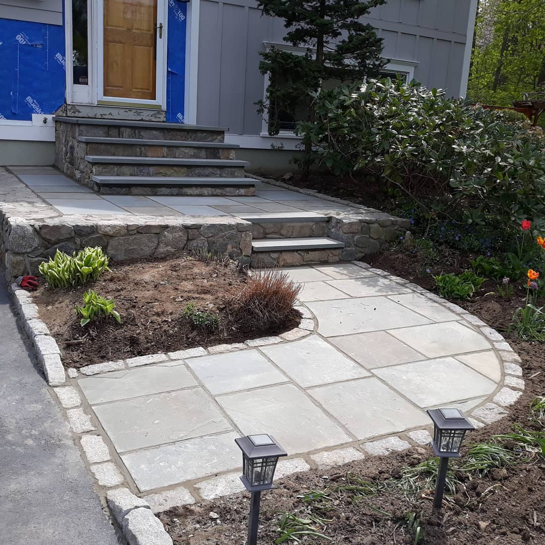 Stone pathway and steps leading to a wooden door, bordered by garden beds and small lantern lights, provide a serene welcome. M Lato ensures that every detail is perfectly crafted, creating an inviting entrance that harmonizes with the surrounding greenery.