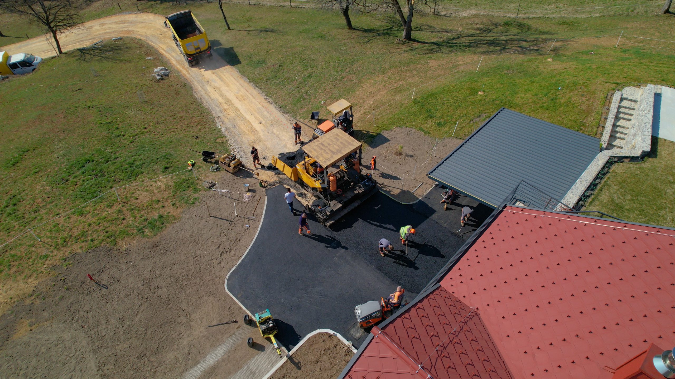 Aerial view of workers from M Lato Excavating & Tree Care Service paving a driveway with asphalt near a house with a red roof, surrounded by lush grass.