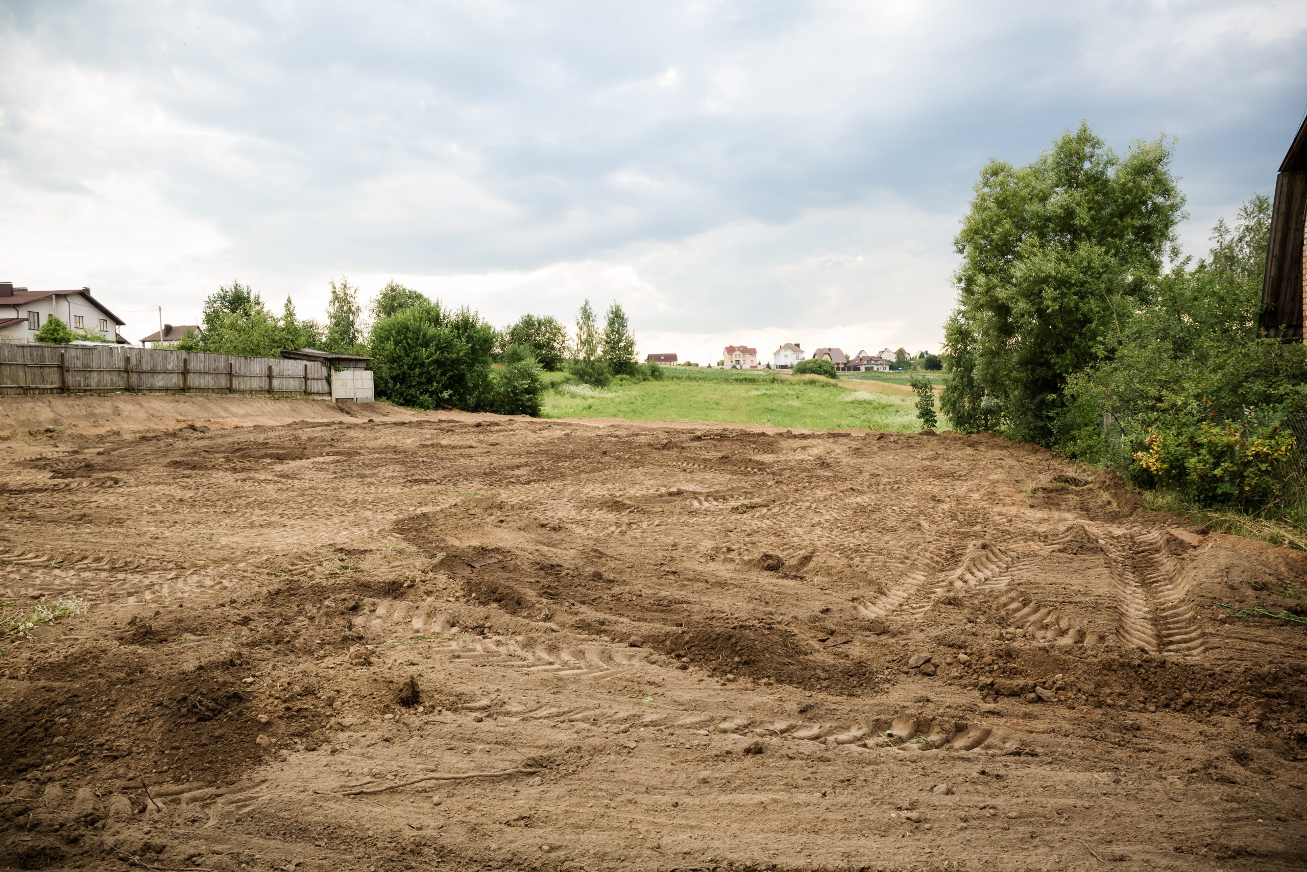 A cleared, dirt-covered plot of land with tire tracks, trees, and houses in the background under a cloudy sky.
