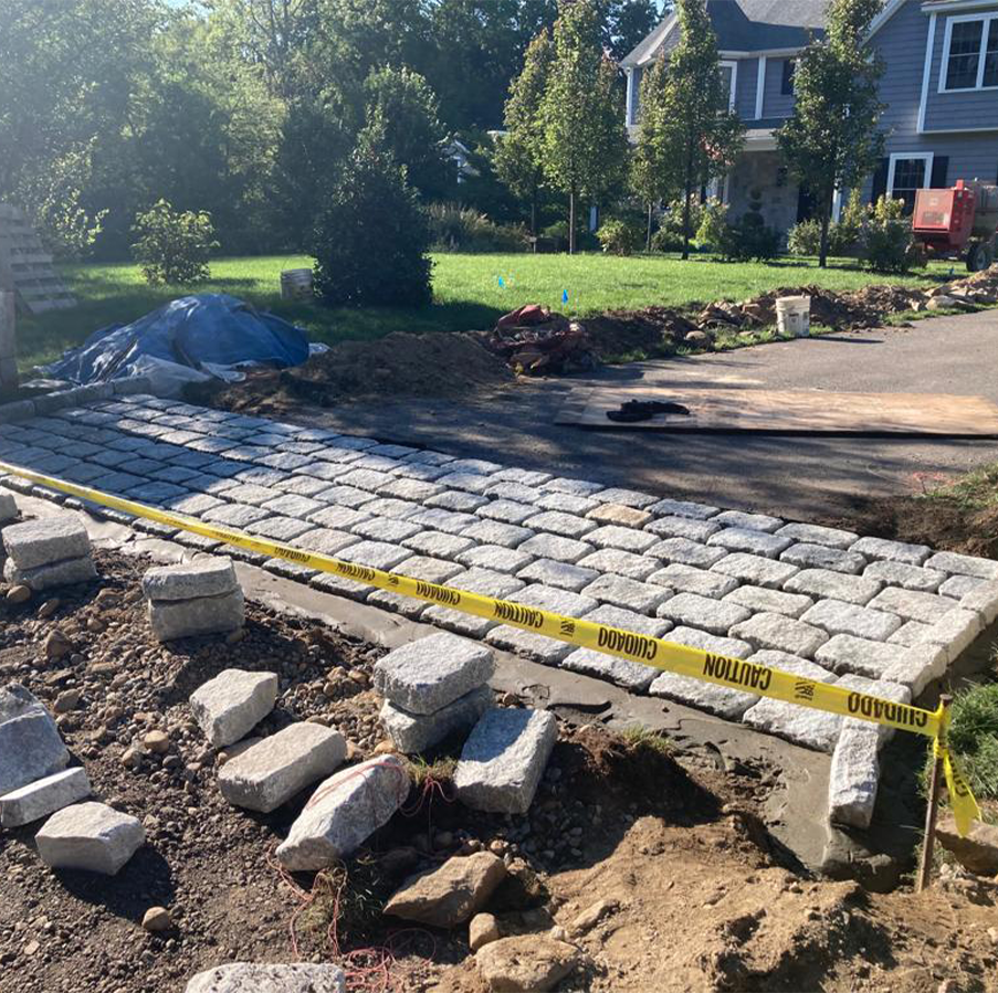 Caution tape blocks a driveway under construction with stone pavers beside a house and yard.