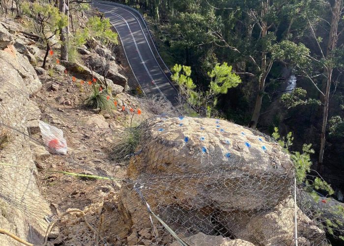 A fenced rock on a hillside near a winding road, with trees and safety markers visible.