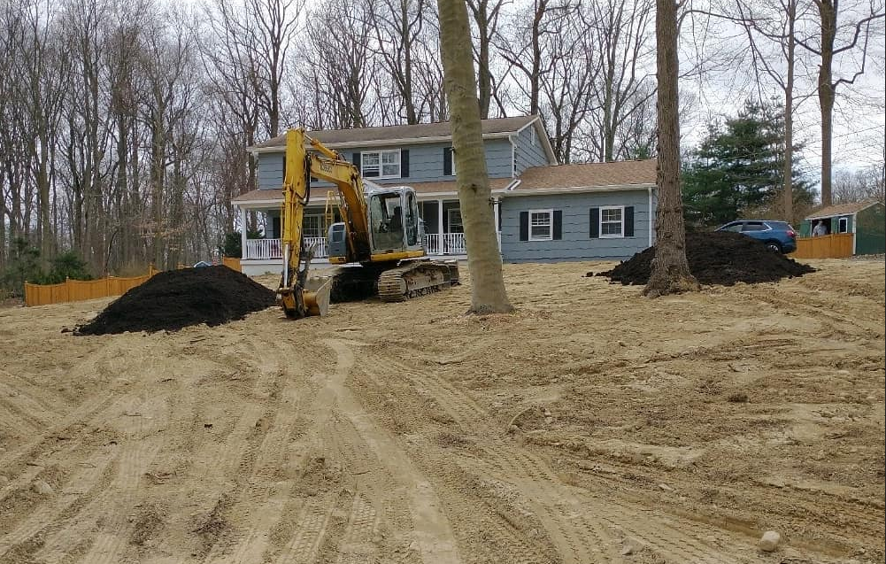 A yellow excavator sits on dirt in front of a house, with piles of soil and leafless trees nearby.