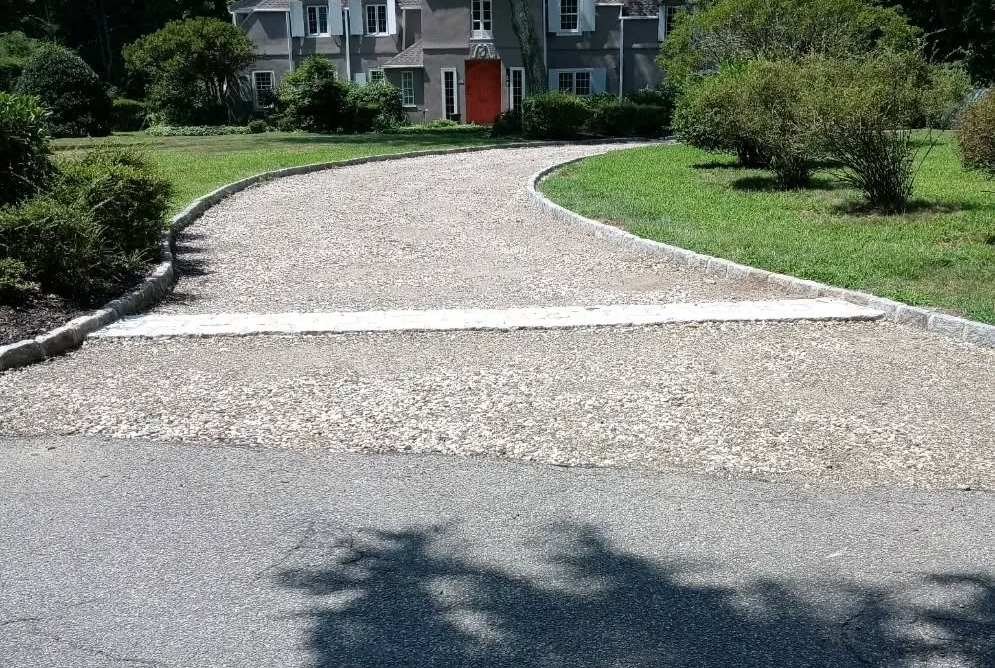 Curved gravel driveway leading to a gray house with a red door, surrounded by bushes and grass.